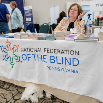 NFB of PA President sits at exhibit hall table. The table has literature and a table covering showing the NFB of PA logo. Lynn's guide dog Danny's head is peaking out from under the table covering.
