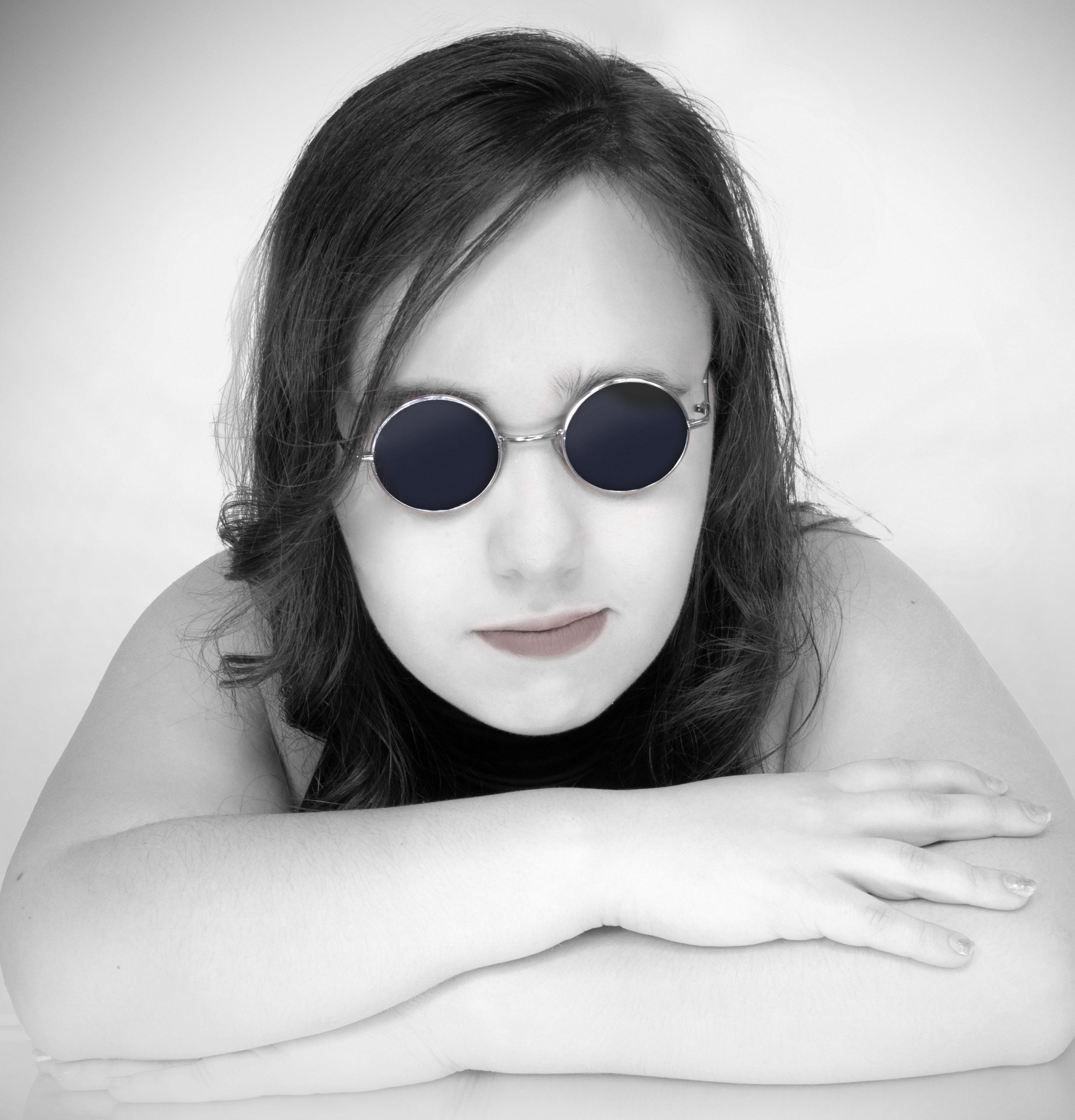 Black and White head shot of Angelina with her chin resting on her arms that are crossed in front of her.  She is wearing John Lennon-style circle sunglasses and a black dress.
